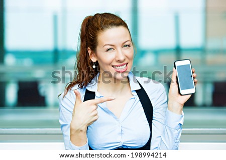 Closeup portrait happy excited, young attractive business woman, company employee showing, holding pointing at her smart phone, isolated background windows corporate office, building. Positive emotion