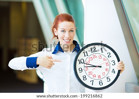 Closeup portrait smiling female, young businesswoman employee manager standing in corporate office hallway pointing on wall clock, isolated background windows. Business, education concept. Punctuality
