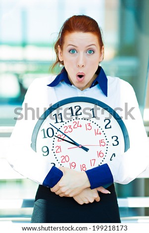 Closeup portrait funny looking, anxious business woman, worker, holding wall clock pressured by lack, running out of time, isolated background corporate office windows. Human face expression, emotion