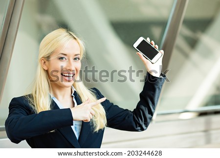 Closeup portrait happy excited, young attractive business woman, company employee showing, holding pointing at her smart phone, isolated background windows corporate office, building. Positive emotion