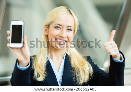 Closeup portrait happy, excited, young, attractive business woman, company employee showing, holding her smart phone, giving thumbs up, isolated background windows corporate office. Positive emotions