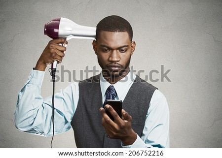 Closeup portrait worried curious business man, deal maker reading text news on smart mobile phone holding hairdryer isolated black grey background. Human face expression emotion of corporate executive