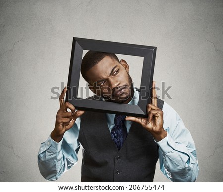 Closeup portrait young businessman executive looking up, curious surprised confused through black picture frame thinking beyond borders accepted rules isolated grey background. Face expression emotion
