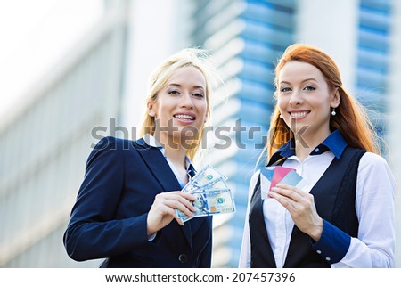 Closeup portrait two smiling business women with credit cards and cash on hands, convenience of electronic money isolated corporate office background. Financial decision, banking system concept