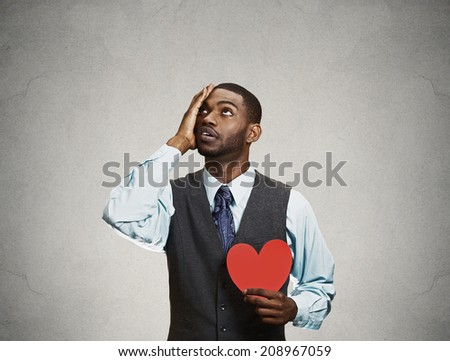 Closeup portrait young sad confused corporate man, holding red heart in his hands, about to cry, looking up isolated grey, black background. Negative human emotion facial expression feelings, attitude