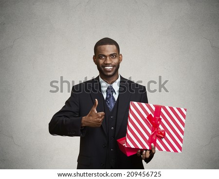 Portrait young happy, smiling business man holding present, red gift box, giving thumb up isolated grey wall background. Positive facial expressions, human emotions, body language, life perception