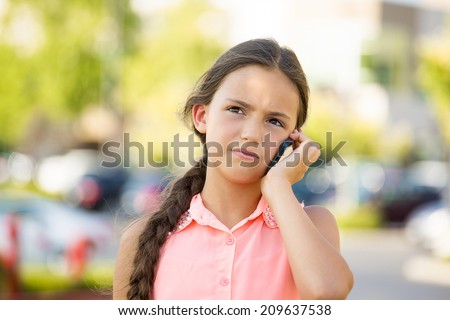 Closeup portrait, worried, sad young child, girl talking on phone to someone, looking unhappy, isolated outdoors background. Negative human emotions, facial expressions, feelings. Communication