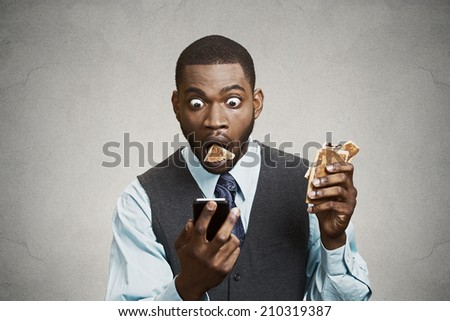 Closeup portrait surprised funny looking corporate business man holding, reading bad news on smart phone while eating cookie, about to choke isolated grey wall background. Face expression, emotion