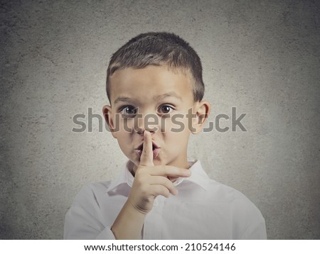 Closeup portrait young serious child, boy placing finger on lips as if to say, shhh, be quiet, silence, isolated grey wall background. Facial expression, human emotions, signs, symbols, body language