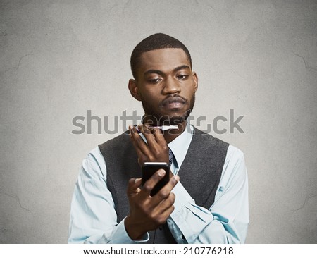 Closeup portrait serious young business man shaving his face, using trimmer and texting, reading news on smart phone, isolated grey wall background. Human face expressions, corporate executive life