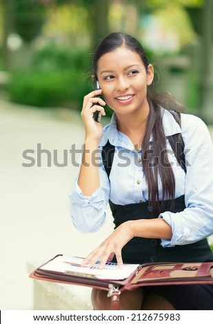 Closeup portrait, beautiful, smiling young business woman in blue shirt, sitting outside, reviewing financial statements, talking on mobile phone isolated park trees background. Facial expressions