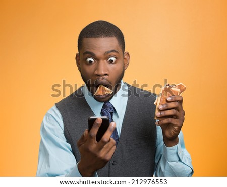 Portrait surprised, funny looking, corporate business man holding, reading bad news on smart phone, eating cookie about to choke isolated orange background. Face expression emotion unexpected reaction