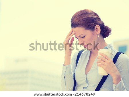 Portrait unhappy young business woman stressed bothered by mistake having bad headache holding cup coffee isolated standing outside corporate office. Negative human emotion expression feeling reaction