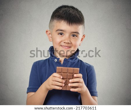 Kids love sweets. Portrait happy little boy eating whole bar of chocolate, isolated on grey wall background. Positive human emotions, face expressions. Food cravings