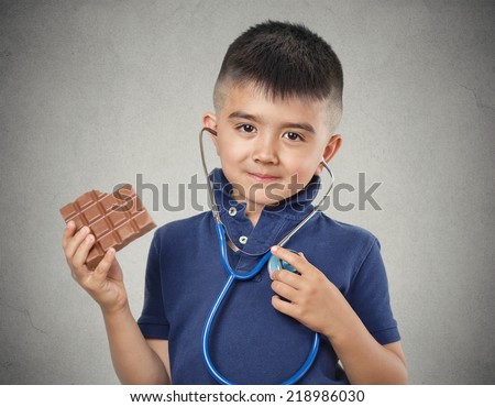 Kids love sweets. Portrait happy little boy eating whole bar of chocolate listening to his heart with stethoscope isolated on grey wall background. Positive human emotion face expression. Food craving