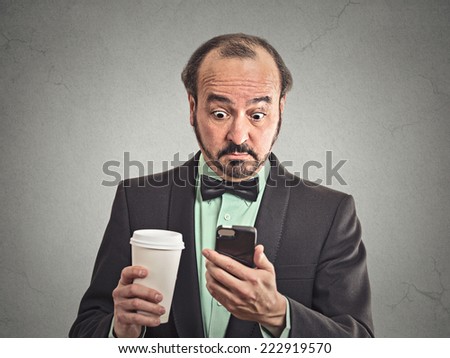 Closeup portrait surprised worried business man reading bad news on smart phone holding mobile drinking cup of coffee isolated grey wall background. Human face expression, corporate executive emotions