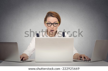 Busy business woman multitasking trying to manage it all by herself working on several computers simultaneously sitting at desk on grey wall office background. Human face expressions, emotions