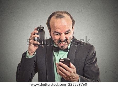 Frustrated angry businessman with alarm clock looking at his smart phone with displeased face expression late for meeting overwhelmed busy schedule isolated grey wall background. Human face expression