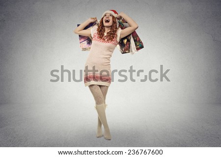 Happy woman in red santa claus hat with christmas gifts waiting fun holidays. Young girl smiling holding shopping bags isolated grey wall background with copy space. Positive face expression