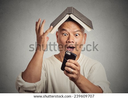 Portrait headshot surprised stunned middle aged man funny looking guy with book on head reading news on smart phone isolated grey wall background. Human face expression emotion reaction perception