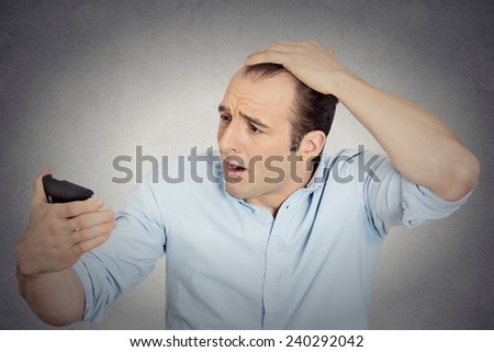 Closeup portrait shocked man feeling head, surprised he is losing hair, receding hairline or seeing bad news on cell phone isolated grey wall background. Negative facial expressions, emotion feeling