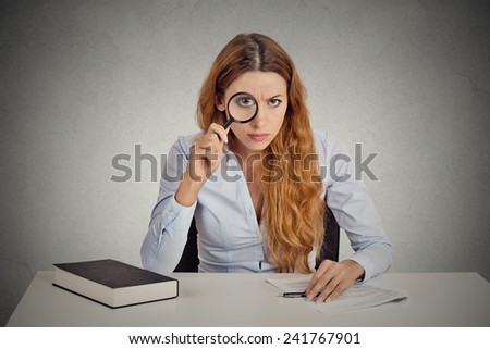 Portrait serious business woman with glasses skeptically looking at you through magnifying glass sitting at desk isolated on office grey wall background. Human face expression, body language, attitude