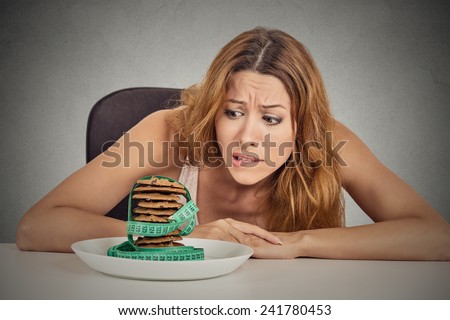 Portrait young unhappy woman craving sugar sweet cookies but worried about weight gain sitting at table isolated grey wall background. Human face expression emotion. Diet nutrition dilemma concept