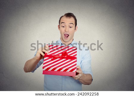 Closeup portrait happy super excited surprised young man about to open unwrap red gift box isolated grey wall background enjoying his present. Positive human emotion facial expression feeling attitude