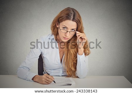 Curious corporate businesswoman with glasses sitting at desk skeptically looking at you scrutinizing isolated on office grey wall background. Human face expression, body language, attitude, perception