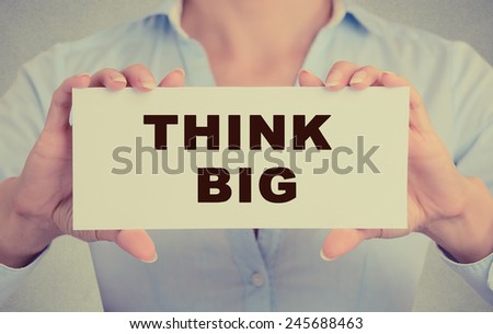 Businesswoman hands holding white card sign with think big text message isolated on grey wall office background. Retro instagram style image