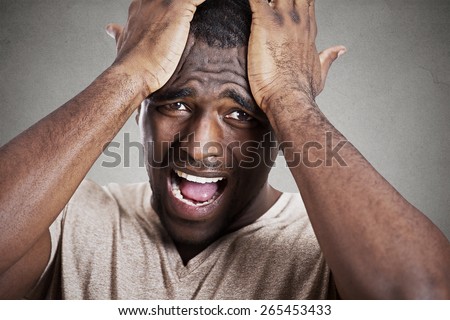 Closeup headshot very sad depressed, stressed disappointed gloomy young man head on hands crying screaming in despair isolated on grey wall background. Human emotion facial expression reaction