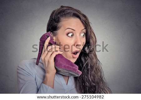 Headshot young surprised woman holding high heeled shoe in her hand as a phone isolated on grey wall background. human face expression emotion feelings reaction