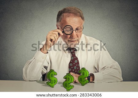 penny pincher. Senior business man looking through magnifying glass at dollar signs symbol on table isolated grey wall background. Economy financial wealth success concept. Ponzi scheme investigation