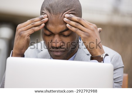 stressed young businessman sitting outside corporate office working on laptop computer holding head with hands looking down. Negative human emotion facial expression feelings.
