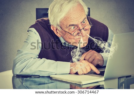 Stressed elderly man using computer blowing steam from nose frustrated sitting at table isolated on gray wall background. Senior people and technology concept