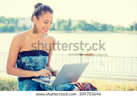 Amused young woman working with her notebook laptop sitting on a lawn by the lake