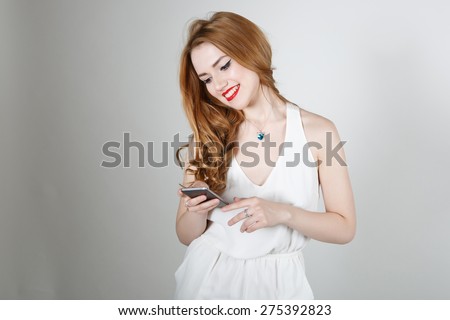 Beautiful girl with red hair and bright red lipstick smiling and looking at the camera on a light background writes sms