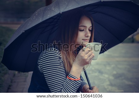 beautiful girl during a rain under umbrella drinks hot coffee outdoor