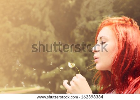beautiful woman with red hair blows into dandelion outdoors