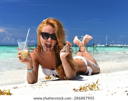 Young happy sexy woman lying relaxing tropical sea beach sand with alcohol cocktail pina colada drink laughing in white bikini and sunglasses