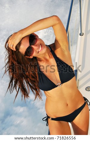 Pretty young slim sexy brunette woman in sunglasses and black stylish bikini on private speed-boat yacht on vacation in summer time smiling and touch her hair, sailing in mediterian sea