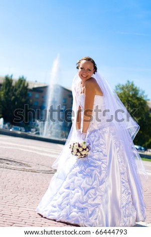 Beautiful bride in white dress holding bouquet and sat next to water fountain.