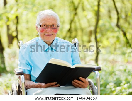 Kind elderly lady in wheelchair with book in hands reading story outdoors.