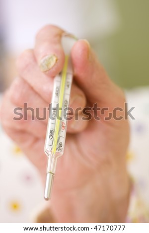 An old woman holding a clinical thermometer in her hand, a blurred image of a doctor in the background.
