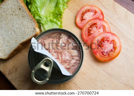 Preparing tuna sandwich, tuna in can opened with bread, lettuce, and tomato on wood background