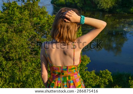 Attractive young lady in summer dress with open back and shoulders standing by the river touching her hair with right hand with blue bracelet.