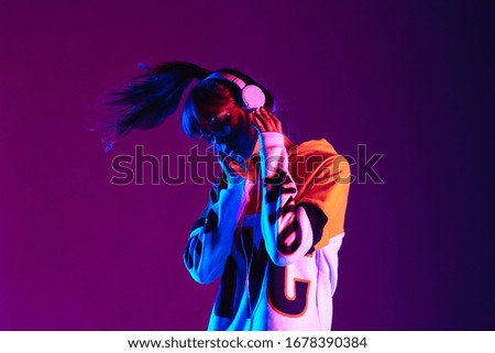 Stylish fashion teenager model wearing hoodie and headphones listening dj music dancing in purple neon lights. Young teen girl enjoy cool music 90s party mix in violet studio background. Copy space.
