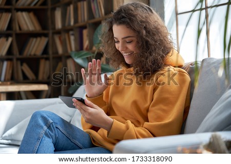 Happy young hispanic latin teen girl sit on sofa at home holding phone looking at screen waving hand video calling distance friend online in mobile chat app using smartphone videochat application.
