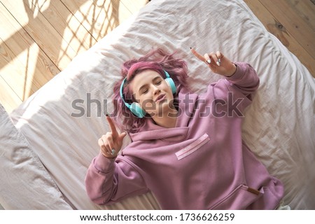Happy funny teen girl with pink hair wear headphones lying in comfortable bed listening new pop music enjoying singing song with eyes closed relaxing in cozy bedroom at home. Top view from above.