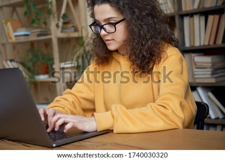 Young hispanic ethnic teen girl college student using laptop computer writing on pc working from home office or studying on tech device in classroom sit at desk. Online education, elearning concept.